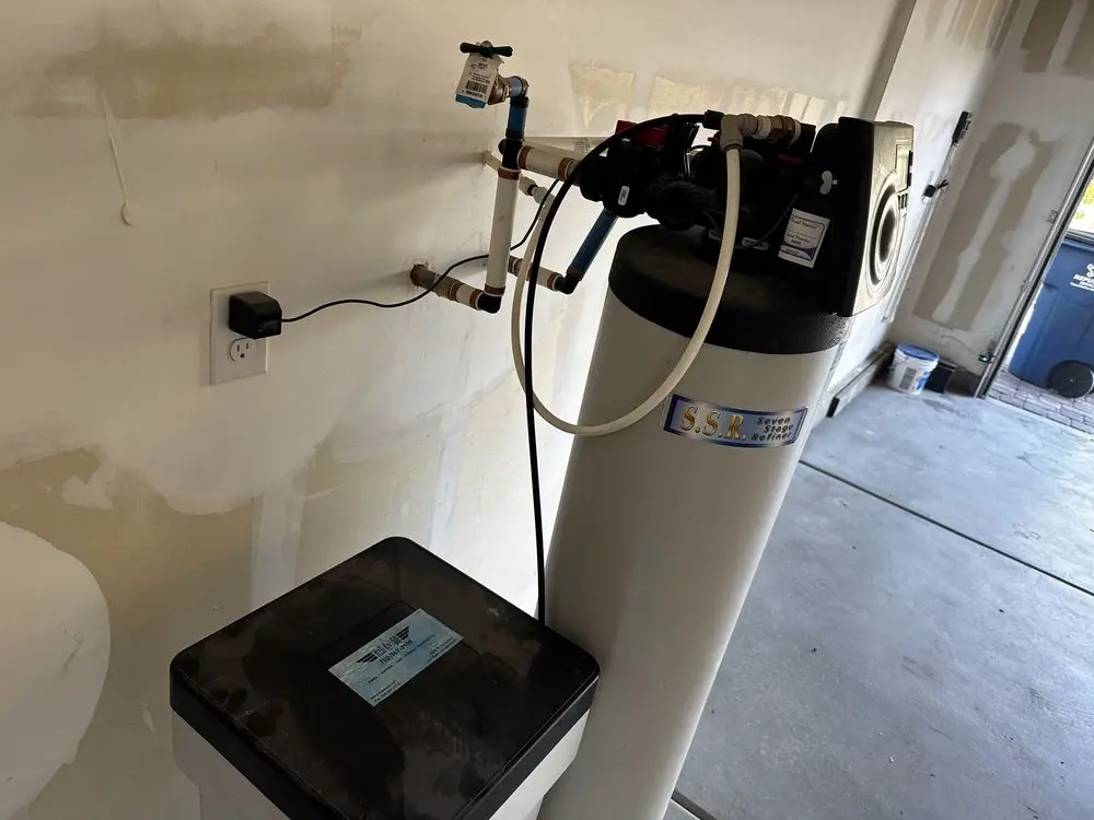 A water treatment system consisting of a tall white cylindrical tank with a control valve and a shorter rectangular tank, installed against a garage wall with plumbing connections - H2O to GO